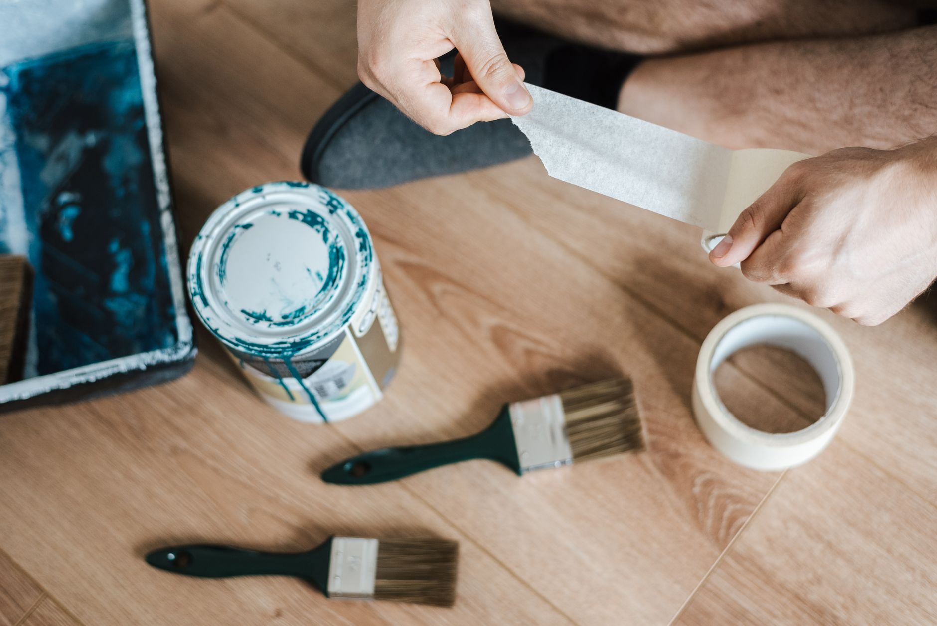 From above of crop anonymous male unwrapping sticky tape roll on parquet near paint brushes in house in Carlsbad, CA